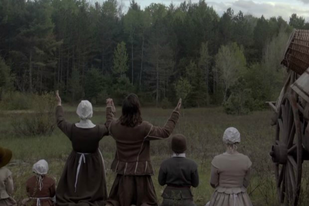 A screenshot from The Witch: A Puritan family kneels before the dark forest and raises their hands in prayer. 