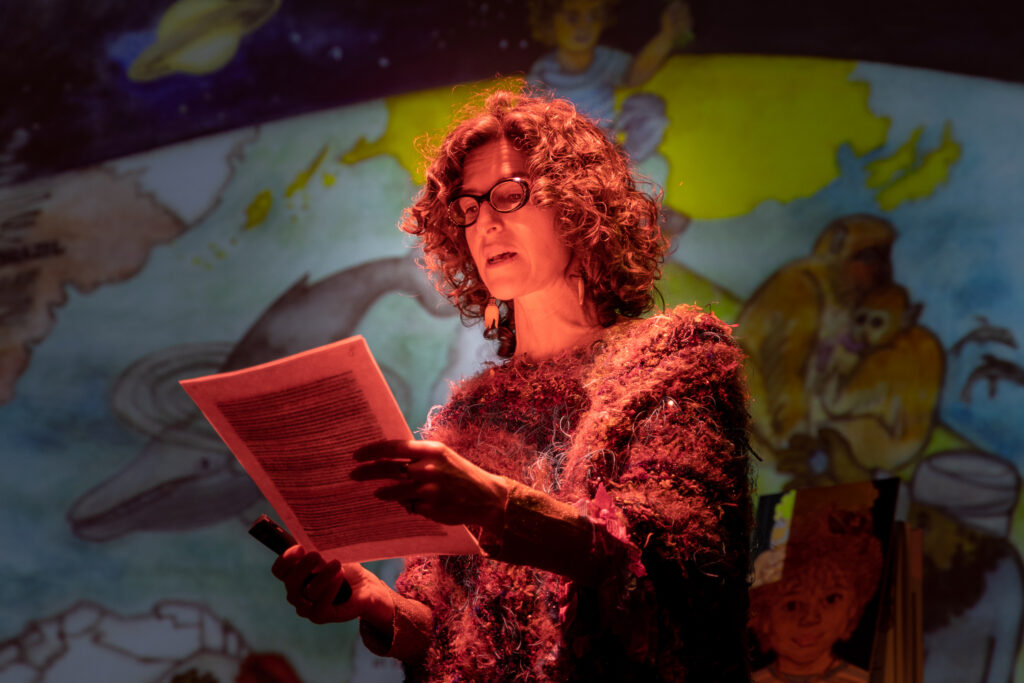 Photo of a woman in red light reading from a paper and speaking.