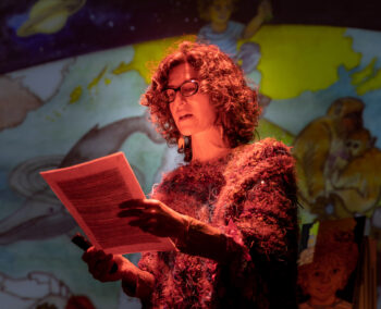 Photo of a woman in red light reading from a paper and speaking.