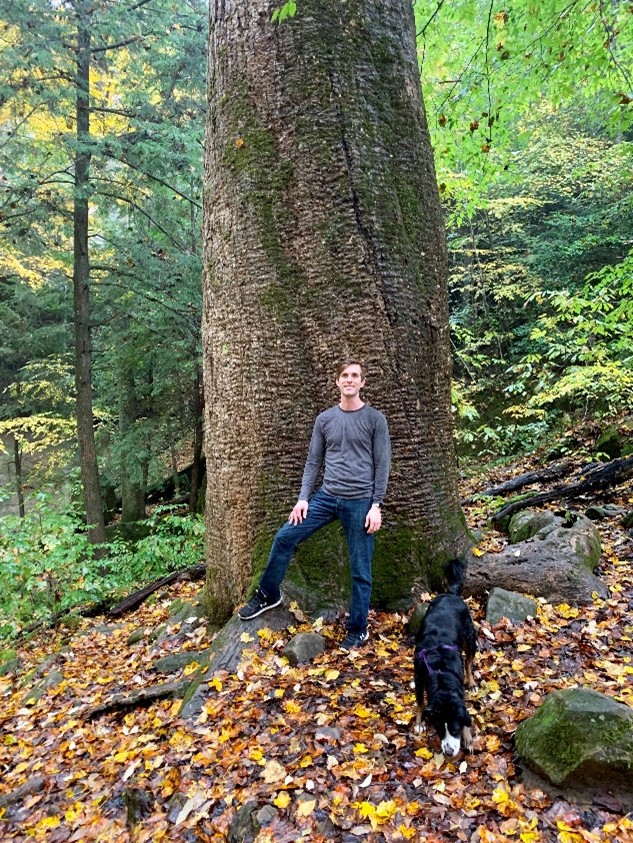 A photo of Nicholas Reich standing in front of the base of a 150-foot tall tree in the forest, with a black dog nearby.